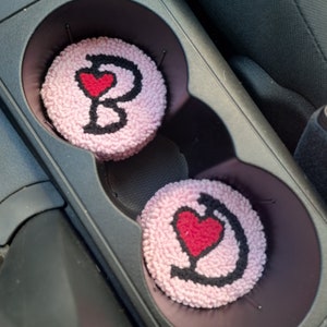 Può includere: Due porta bicchieri per auto rosa con ricami neri e rossi. Un porta bicchieri ha un cuore e la lettera "C" ricamata. L'altro porta bicchieri ha un cuore e la lettera "B" ricamata.