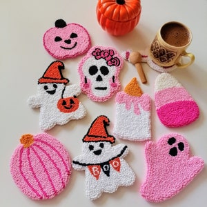 May include: A set of ten Halloween-themed punch needle coasters. The coasters are made of pink and white yarn and feature various designs, including pumpkins, ghosts, a skull, and a candy corn.
