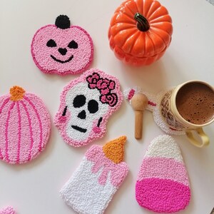 May include: Six pink and white crocheted coasters with various Halloween-themed designs, including a pumpkin, a skull, a candle, and a candy corn. The coasters are arranged on a white surface with a ceramic pumpkin and a cup of coffee.