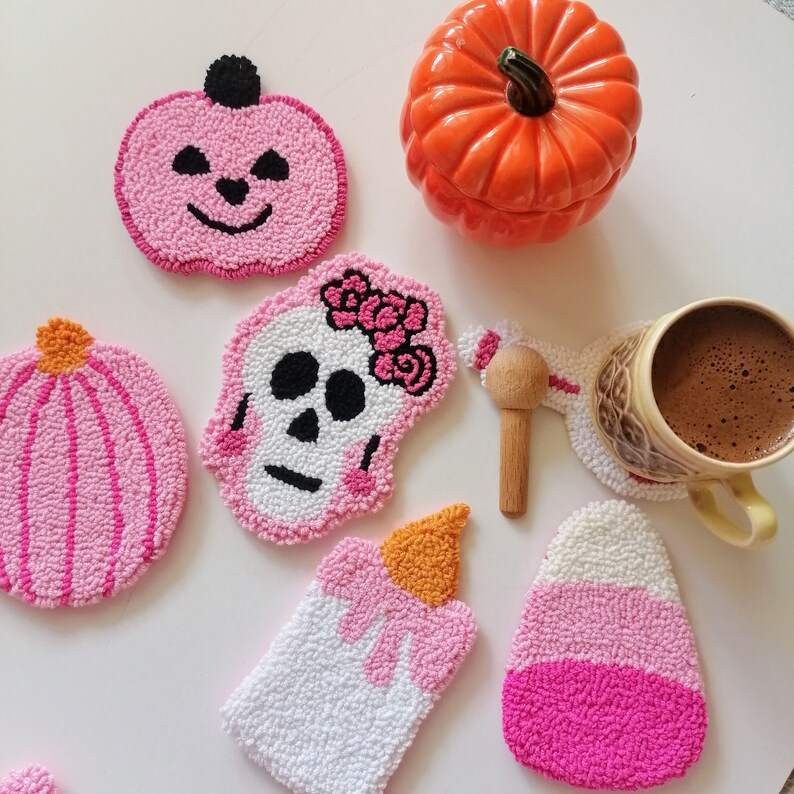 May include: Six pink and white crocheted coasters with various Halloween-themed designs, including a pumpkin, a skull, a candle, and a candy corn. The coasters are arranged on a white surface with a ceramic pumpkin and a cup of coffee.