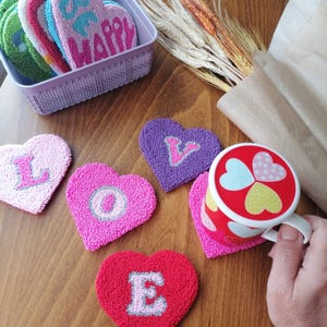 May include: A set of six heart-shaped coasters with the letters "LOVE" embroidered in pink, purple, and red. The coasters are made of a soft, plush material and are perfect for adding a touch of whimsy to your home decor.