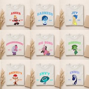 May include: Nine light-colored t-shirts, each featuring a different emotion from the movie "Inside Out." The shirts display the words "ANGER," "SADNESS," "JOY," "EMBARRASSMENT," "BIG BONG," "DISGUST," "ANXIETY," "ENVY," and "ENNUI" with corresponding character illustrations.