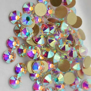 May include: A close-up of iridescent rhinestones and gold-coloured, flat-backed circles. The rhinestones display a range of colours, including pink, purple, and green. These embellishments are suitable for various craft projects.