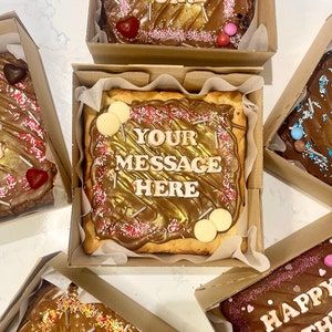 May include: Assortment of decorated brownie squares in cardboard boxes. The brownies are topped with chocolate, sprinkles, and gold lettering. One brownie says "Your message here", another says "Girl", and another says "Happy Birthday".