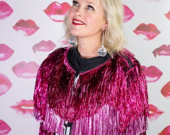 May include: A woman wearing a pink sequined jacket with fringe, a black t-shirt with the word "ALL" on it, and black jeans. She is standing in front of a white background with pink lips painted on it.