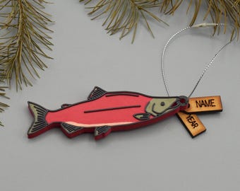 Puede incluir: Un adorno de pez rojo de madera con detalles en negro y beige, y una cuerda plateada para colgar. Se adjuntan dos pequeñas etiquetas de madera, una con "NAME" y otra con "YEAR". El adorno está cerca de agujas de pino.