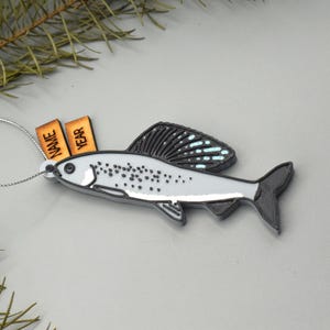 Puede incluir: Adorno navideño con forma de pez, con una cuerda plateada para colgar. El adorno es principalmente gris con detalles en negro y una aleta negra. Dos pequeñas etiquetas de madera dicen "NAME" y "YEAR".