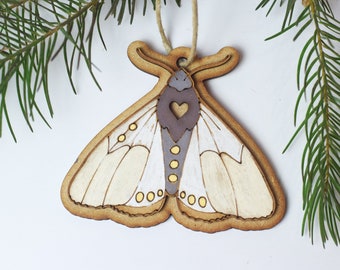May include: A wooden ornament shaped like a moth with white wings and a gray body. The moth has a heart-shaped cutout in its body and yellow dots on its wings.