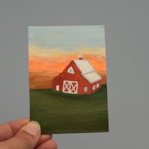 Puede incluir: Pequeña pintura de un granero rojo con techo y detalles blancos, sobre un cielo de atardecer. El granero presenta una puerta blanca y un diseño en forma de cruz. La obra de arte está sobre un papel rectangular.