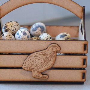 Puede incluir: Una caja de madera con asa, llena de huevos de codorniz moteados. Una peque&ntilde;a codorniz est&aacute; tallada en la parte delantera de la caja.