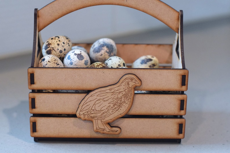 Puede incluir: Una caja de madera con asa, llena de huevos de codorniz moteados. Una peque&ntilde;a codorniz est&aacute; tallada en la parte delantera de la caja.