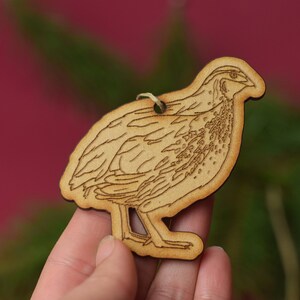 May include: A wooden ornament shaped like a quail, with a detailed outline of the bird's feathers. The ornament is light brown and has a hole at the top for hanging.