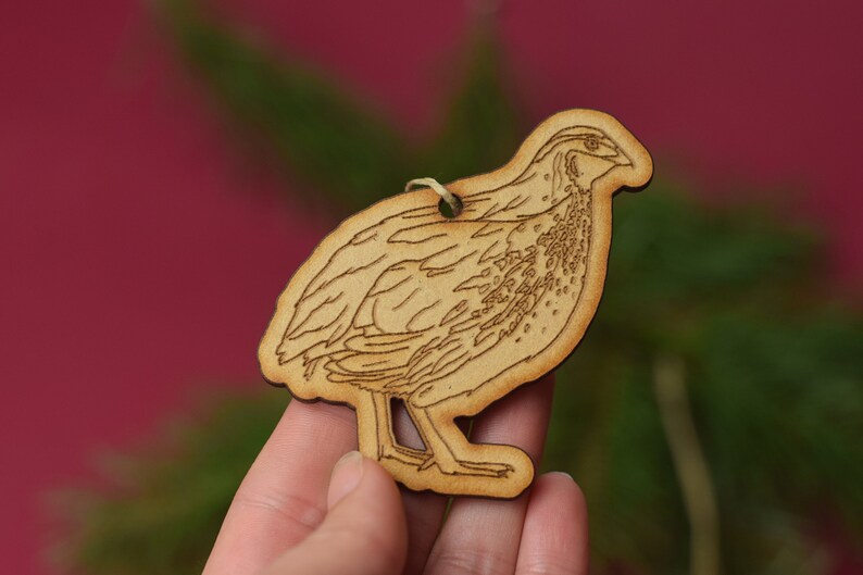 May include: A wooden ornament shaped like a quail, with a detailed outline of the bird's feathers. The ornament is light brown and has a hole at the top for hanging.