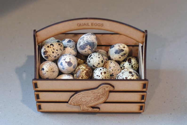 Puede incluir: Una caja de madera con asa, etiquetada con "QUAIL EGGS", llena de huevos de codorniz moteados. La caja tiene un recorte de una codorniz en la parte delantera.