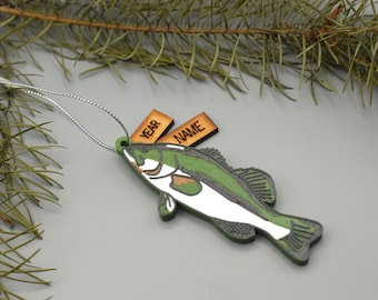 May include: A wooden fish ornament with green, white, and gray details, and two small wooden tags that read "YEAR" and "NAME". The ornament is hanging by a silver string, and is set against a background of pine needles.