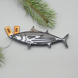 May include: A fish-shaped Christmas ornament with a gray, white, and black design. The ornament has a silver string for hanging and two small wooden tags that read "NAME" and "YEAR". A sprig of evergreen is in the background.