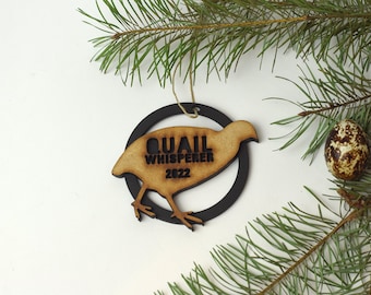 May include: A wooden Christmas ornament shaped like a quail with the text "Quail Whisperer 2022" on it. The ornament is hanging from a string and is surrounded by pine branches.