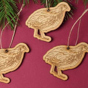 May include: Three wooden ornaments shaped like quail, each with a brown twine string for hanging. The ornaments are on a burgundy background.