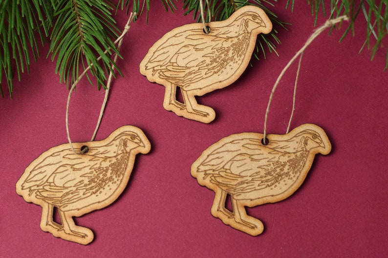May include: Three wooden ornaments shaped like quail, each with a brown twine string for hanging. The ornaments are on a burgundy background.