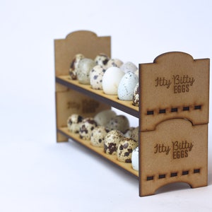 May include: A two-tiered wooden egg holder with the text "Itty Bitty EGGS" on the side. The holder is filled with small, speckled quail eggs.