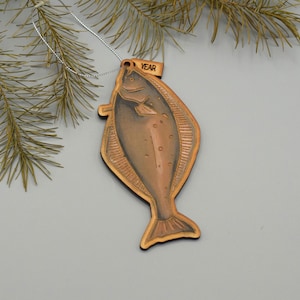 May include: A wooden fish-shaped Christmas ornament with a copper finish. The ornament has a small tag that reads "YEAR". It hangs from a silver string, with a pine branch in the background. The fish is a flat shape.