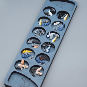 Könnte beinhalten: Ein blaues Mancala-Spielbrett aus Holz mit zwölf kreisförmigen Vertiefungen, die jeweils mit kleinen, farbigen Steinen gefüllt sind. Die Steine sind in verschiedenen Farben gehalten, darunter Weiß, Gelb, Blau und Schwarz. Das Brett hat ein Halbmond-Design.