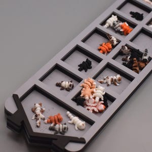 May include: A long, rectangular gray tray with multiple square compartments containing numerous miniature cat figurines in various colors including white, black, orange, and brown. The tray features a dark gray border and a pointed end design.