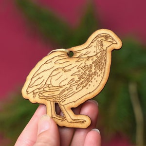 May include: A wooden ornament shaped like a quail, with a detailed laser-cut design. The ornament is light brown and has a hole at the top for hanging.