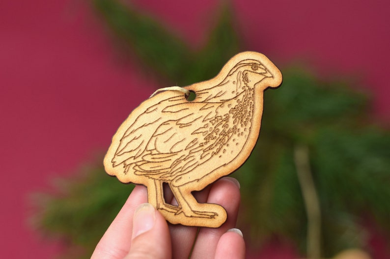 May include: A wooden ornament shaped like a quail, with a detailed laser-cut design. The ornament is light brown and has a hole at the top for hanging.