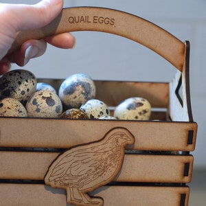 Puede incluir: Una caja de madera con asa, etiquetada con "QUAIL EGGS", llena de huevos de codorniz moteados. Una peque&ntilde;a silueta de codorniz cortada con l&aacute;ser se encuentra en la parte delantera de la caja.