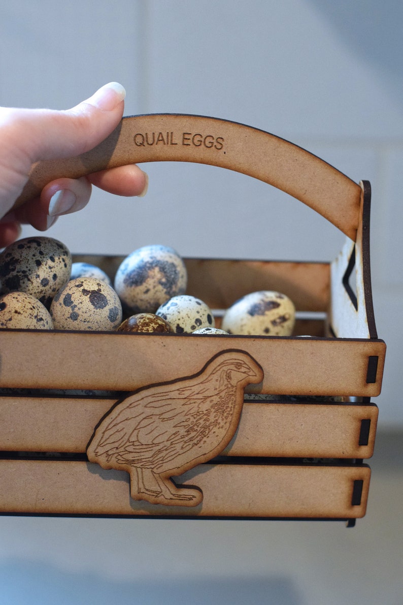 Puede incluir: Una caja de madera con asa, etiquetada con "QUAIL EGGS", llena de huevos de codorniz moteados. Una peque&ntilde;a silueta de codorniz cortada con l&aacute;ser se encuentra en la parte delantera de la caja.