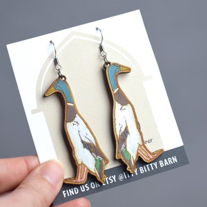 Könnte beinhalten: Ein Paar hölzerne Entenohrringe mit bemalten Details. Die Enten sind braun, weiß und blau mit gelben Schnäbeln. Die Ohrringe hängen an silbernen Haken. Die Karte lautet "Find us on Etsy @ Lil' Bitty Barn".