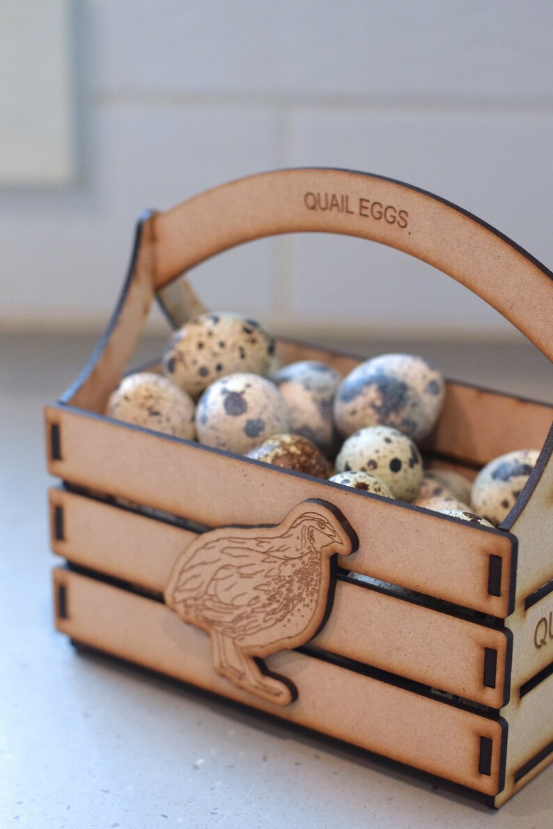 Puede incluir: Una caja de madera con asa, etiquetada con "QUAIL EGGS", llena de huevos de codorniz moteados. La caja tiene un recorte de una codorniz en la parte delantera.