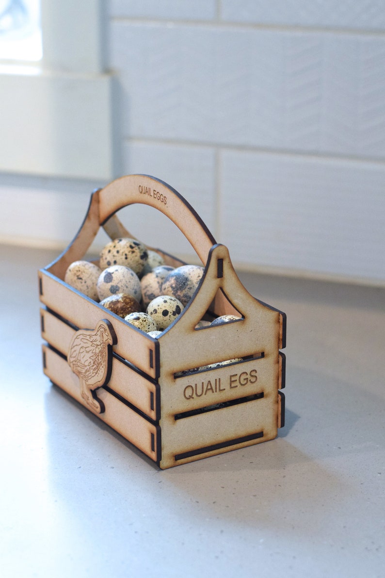 Puede incluir: Una caja de madera con asa, etiquetada con "Quail Eggs", llena de huevos de codorniz moteados. La caja tiene un recorte de una codorniz en el lateral.