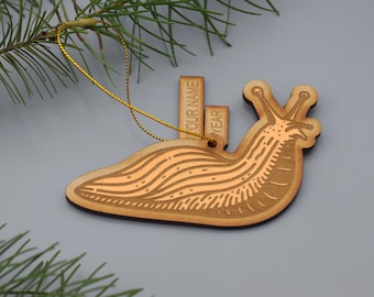May include: A wooden Christmas ornament shaped like a snail with a gold string for hanging. The ornament has the words "OUR NAME" and "YEAR" printed on it.