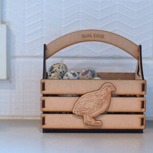 Puede incluir: Una caja de madera con asa, etiquetada como "QUAIL EGGS", contiene una colecci&oacute;n de huevos de codorniz moteados. Un recorte de una codorniz est&aacute; en la parte delantera de la caja.