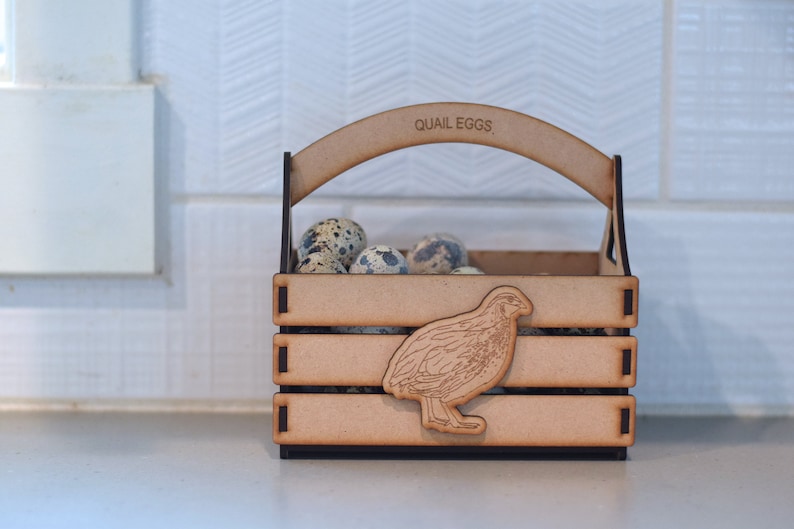 Puede incluir: Una caja de madera con asa, etiquetada como "QUAIL EGGS", contiene una colecci&oacute;n de huevos de codorniz moteados. Un recorte de una codorniz est&aacute; en la parte delantera de la caja.