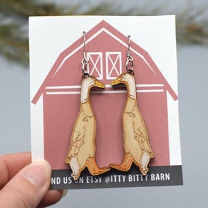 Könnte beinhalten: Holz-Ohrringe mit Enten-Motiv und silberfarbenen Haken. Die Ohrringe zeigen eine detaillierte Illustration einer Ente mit cremefarbenem Körper, weißem Hals und orangefarbenem Schnabel und Füßen. Auf einer weißen Karte mit roter Scheunen-Grafik präsentiert.