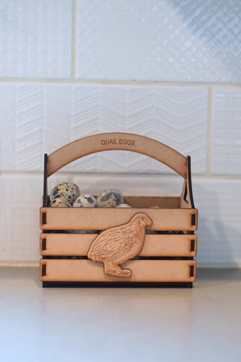Puede incluir: Una caja de madera con asa, etiquetada con "QUAIL EGGS", llena de huevos de codorniz moteados. Una peque&ntilde;a ilustraci&oacute;n de codorniz est&aacute; en la parte delantera de la caja.