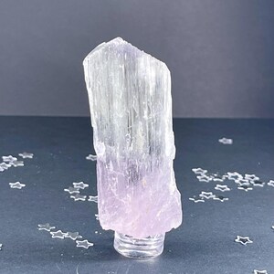 May include: A large, clear, and translucent crystal with a pink hue at the base. The crystal is on a clear stand. The crystal has a striated texture.