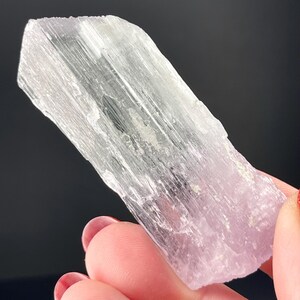 May include: A translucent, light pink and white crystal with a striated texture. The crystal is shaped like a long, thin prism.