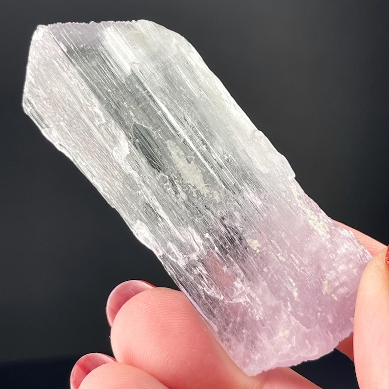 May include: A translucent, light pink and white crystal with a striated texture. The crystal is shaped like a long, thin prism.