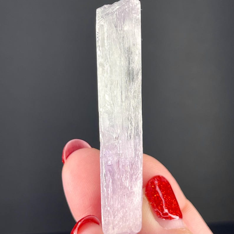 May include: A translucent, white, rectangular crystal with a striated texture. The crystal is approximately 5 inches long and 1 inch wide.