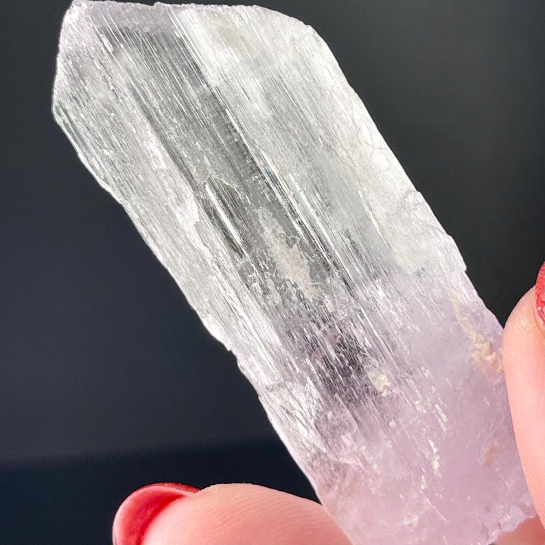 May include: A clear, translucent, elongated crystal with striations. The crystal is a light shade of pink and has a smooth, polished surface.