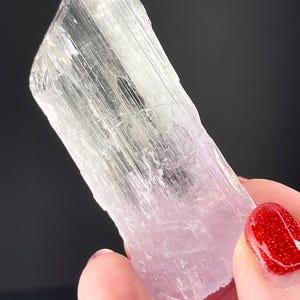 May include: A clear, translucent, elongated crystal with a pale pink hue and striations. The crystal is held in a hand with red nail polish.