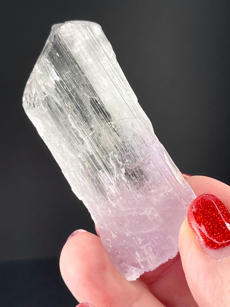 May include: A clear, translucent, elongated crystal with a pale pink hue and striations. The crystal is held in a hand with red nail polish.
