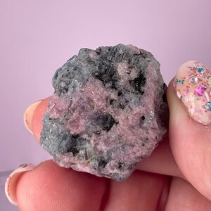 May include: A rough, raw, pink and grey gemstone with a natural, irregular shape. The stone has a mix of pink and grey hues, with some darker grey areas.