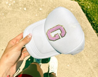 May include: White trucker hat with a pink and gold letter G embroidered on the front.