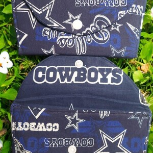 May include: Two navy blue fabric wallets with white and blue star and text patterns. The top wallet is closed with a white button. The bottom wallet is open, revealing the interior. The word "COWBOYS" is visible on the bottom wallet.