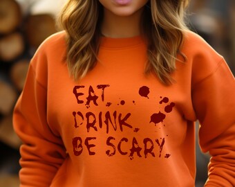 May include: An orange crewneck sweatshirt featuring the dark red text EAT, DRINK, BE SCARY, accompanied by several dark red splatters resembling blood drops on the front of the garment. The sweatshirt has long sleeves and a ribbed neckline.